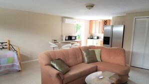 Ein Wohnzimmer mit einer Couch, einem Glastisch, einem gemusterten Teppich, einem Bett mit farbenfroher Decke und einer Küchenzeile im Hintergrund.