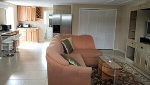 Ein Wohnzimmer mit Sofa, Couchtisch und TV-Schrank. Im Hintergrund ist eine Küchenzeile mit Schränken und Geräten zu sehen.