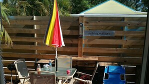 Eine Holzterrasse mit Tisch und Stühlen, einem blauen Liegestuhl und einem bunten Sonnenschirm.