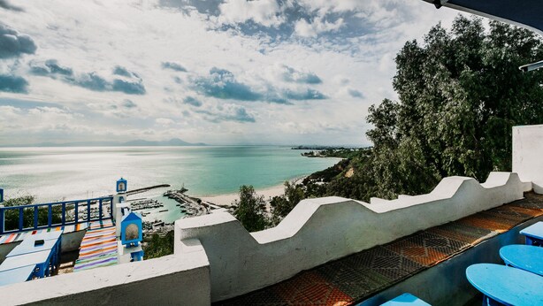 Une terrasse en bord de mer offrant une vue sur la mer et le paysage environnant, avec une barrière en béton, des tables bleues et un tapis rayé aux couleurs vives.