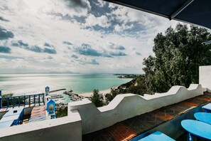 Eine Strandterrasse mit Blick auf das Meer und die Ferne, mit einer Betonbarriere, blauen Tischen und einer bunten, gestreiften Matte.