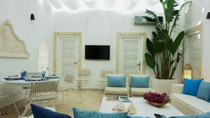 Un salon moderne avec un canapé blanc, des coussins bleus, une table basse avec des livres et un vase de fleurs, un téléviseur fixé au mur et une grande plante.