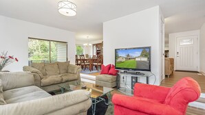 Una sala de estar moderna con un sofá color beige, un sillón rojo y una mesa de centro de vidrio. Un televisor de pantalla plana está montado en la pared, y se distingue una zona de comedor al fondo.