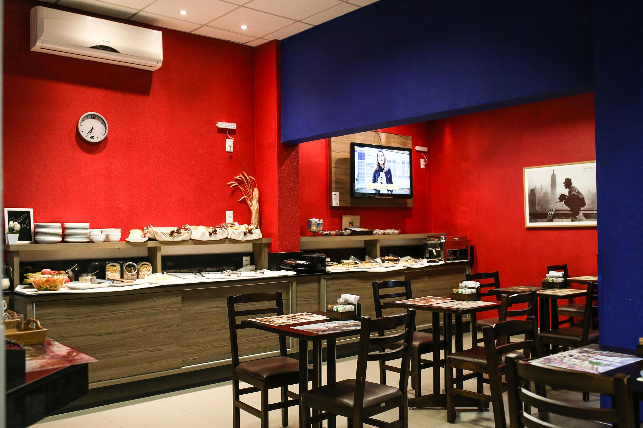 Un restaurante con una decoración en tonos rojo y azul, una barra de buffet y una pantalla de televisión que muestra un noticiero.