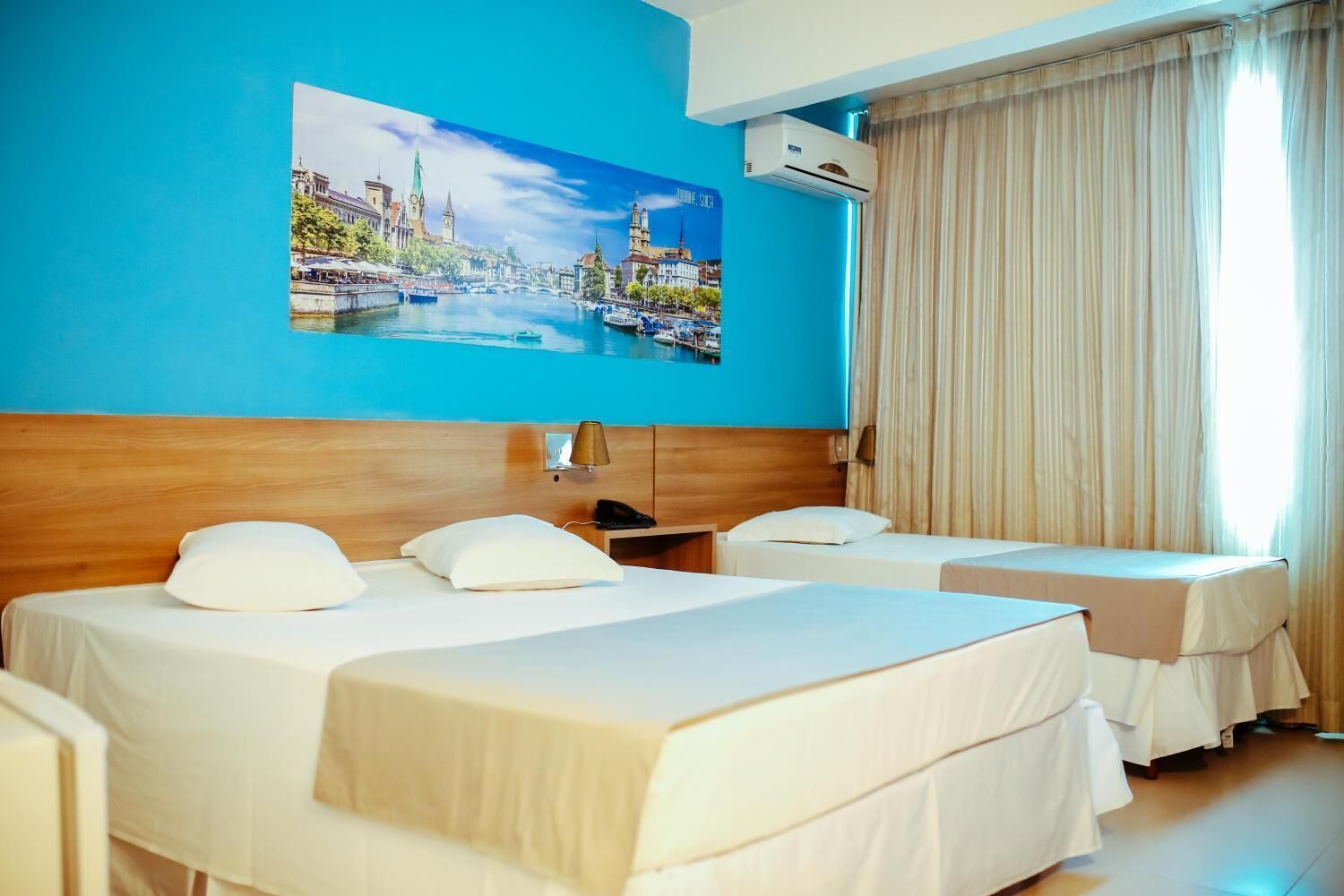 Habitación de hotel con dos camas, un ventanal con cortinas, aire acondicionado montado en la pared y un cuadro con un paisaje urbano.