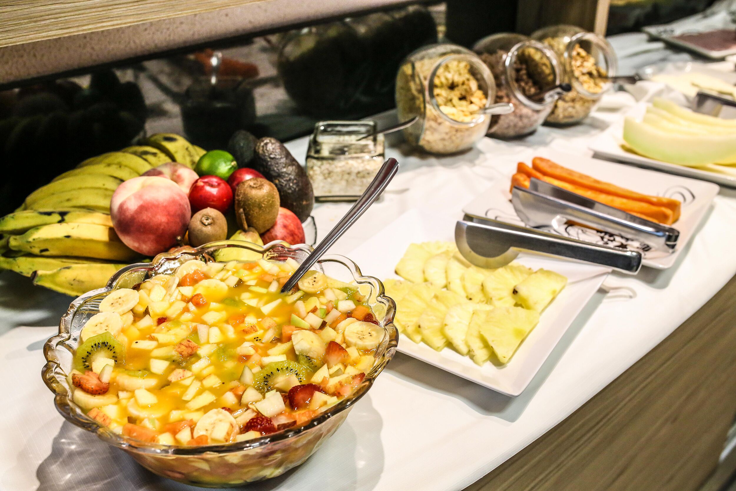 Buffet com diversas frutas, uma saladeira de fruta e vários recipientes com comida.