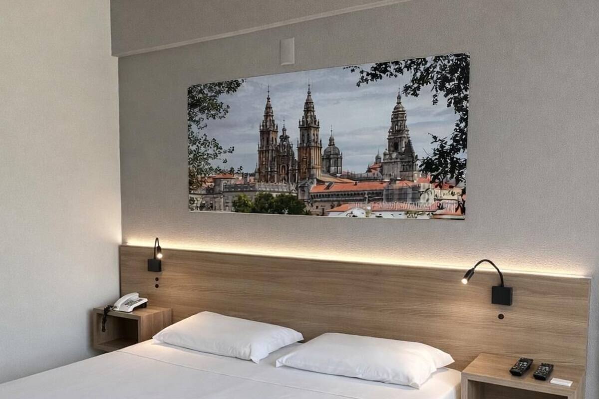 A modern hotel room with a large bed, two bedside tables, a nightstand, a wall-mounted lamp, and a framed cityscape picture.