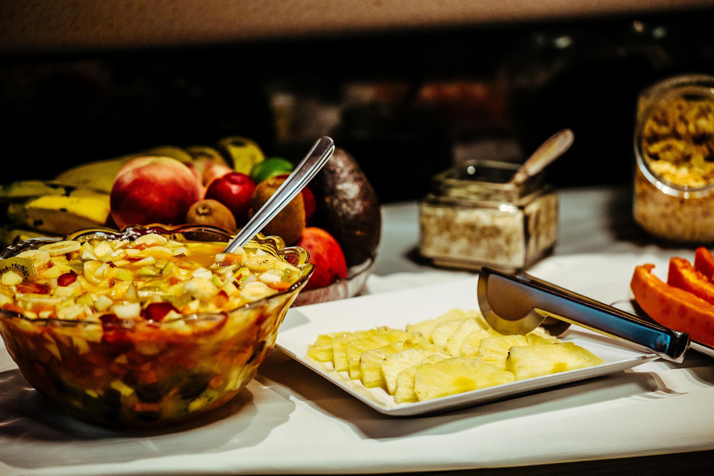 Una mesa de buffet con diversos alimentos, incluyendo un bol de frutas, un plato de rodajas de piña y un frasco que parece contener un postre o salsa.