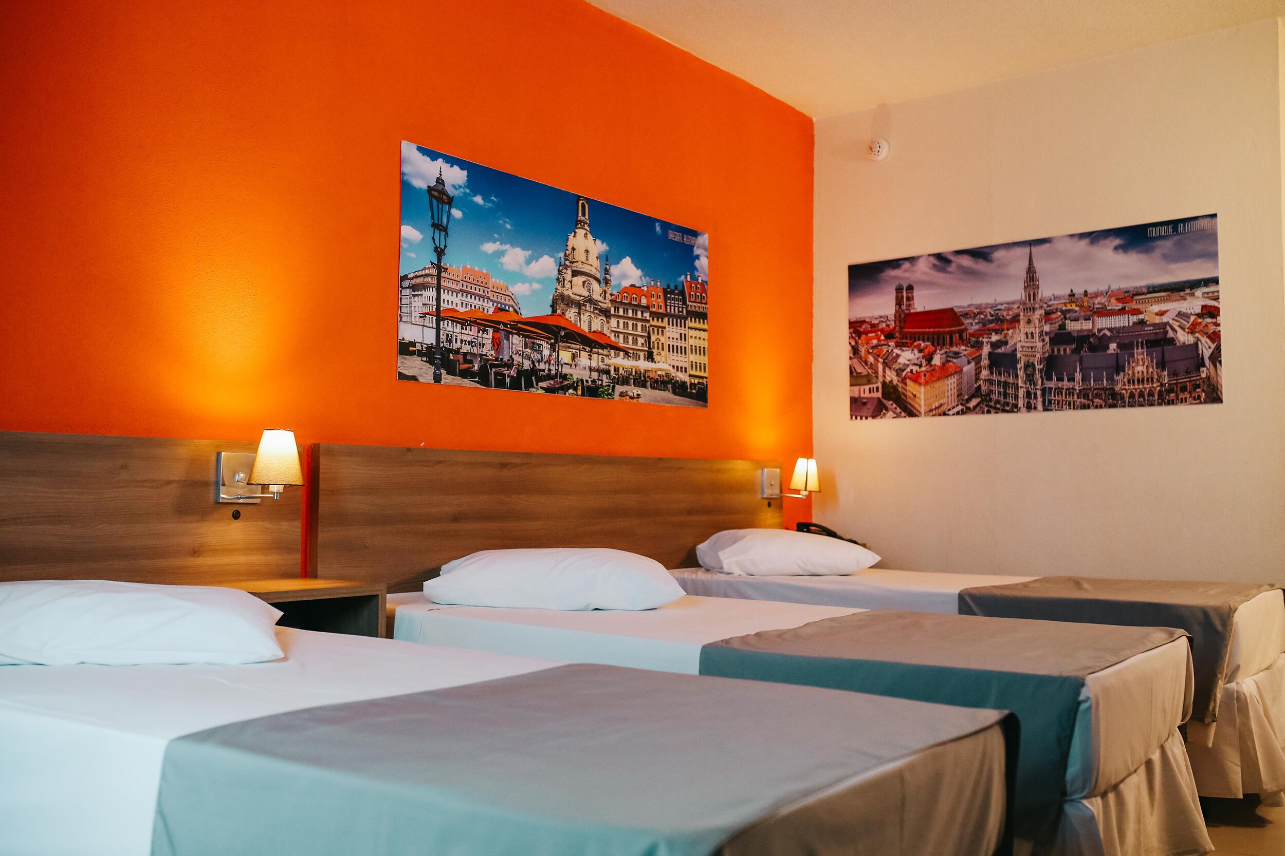 Habitación de hotel con tres camas, paredes anaranjadas y dos cuadros con vistas urbanas.