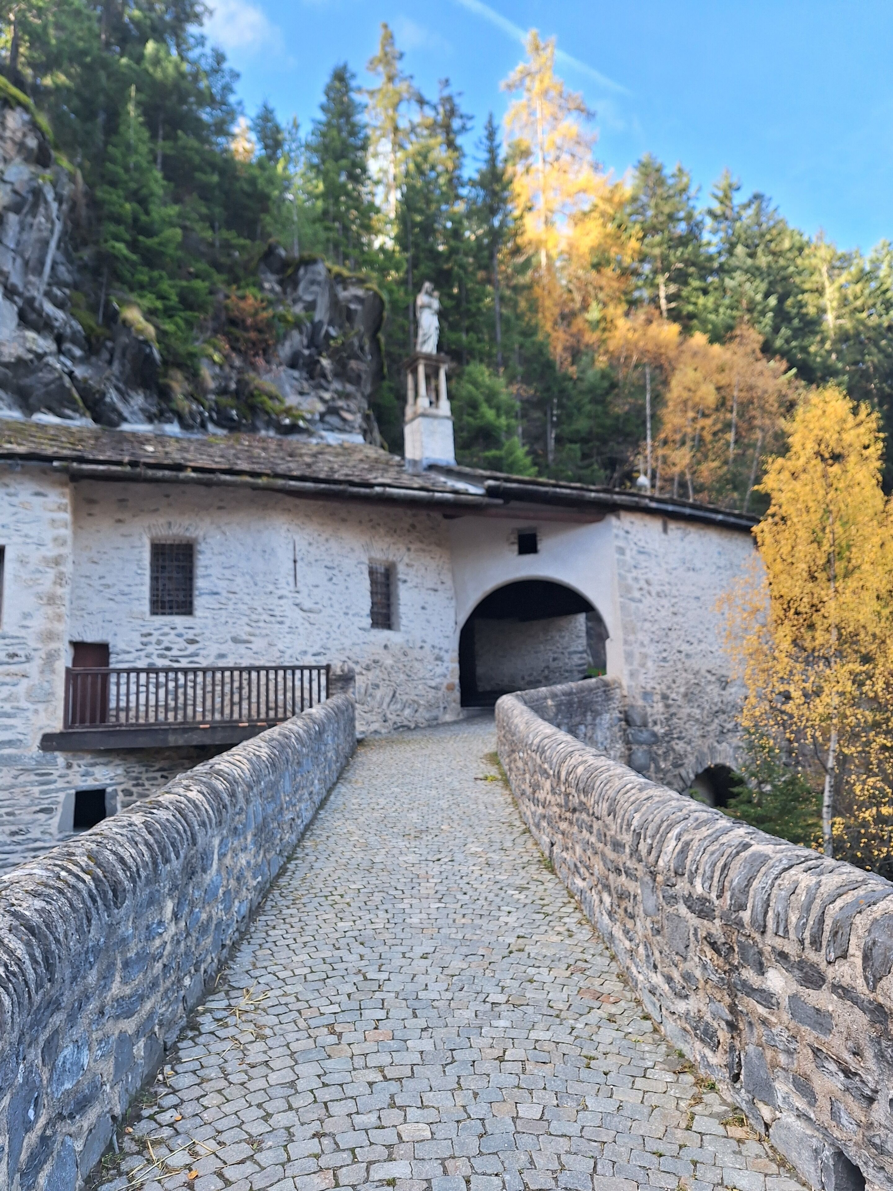 A stone pathway leading to a rustic building with a statue on the roof, surrounded by autumn trees.