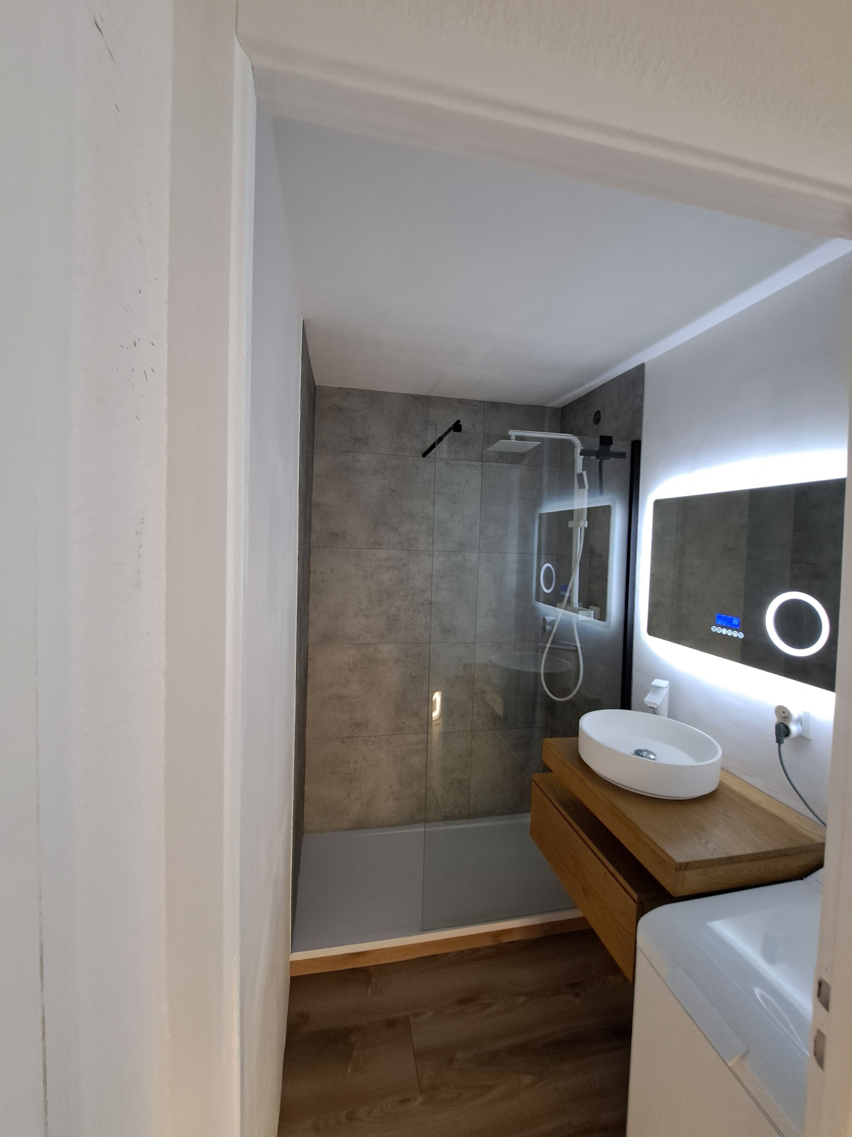 Un bagno moderno con cabina doccia in vetro, un mobile bagno in legno con lavandino bianco e uno specchio con illuminazione integrata.