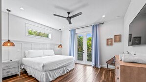 A modern bedroom with a large bed, wooden flooring, a ceiling fan, and a view of greenery through the window.