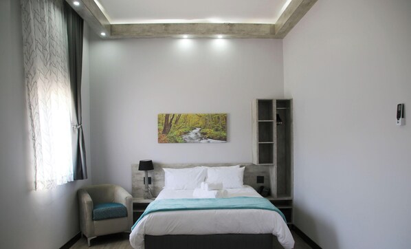 A modern hotel room with a bed, a chair, a painting, a nightstand, and a wardrobe. - King's Gate Hotel Rustenburg (Rustenburg)