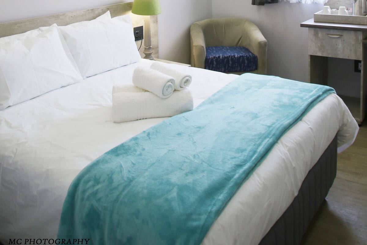 A neatly made bed with white linens and a teal throw blanket, a green lamp beside the bed, a chair, a desk with a mirror, and a painting of a waterfall on the wall.