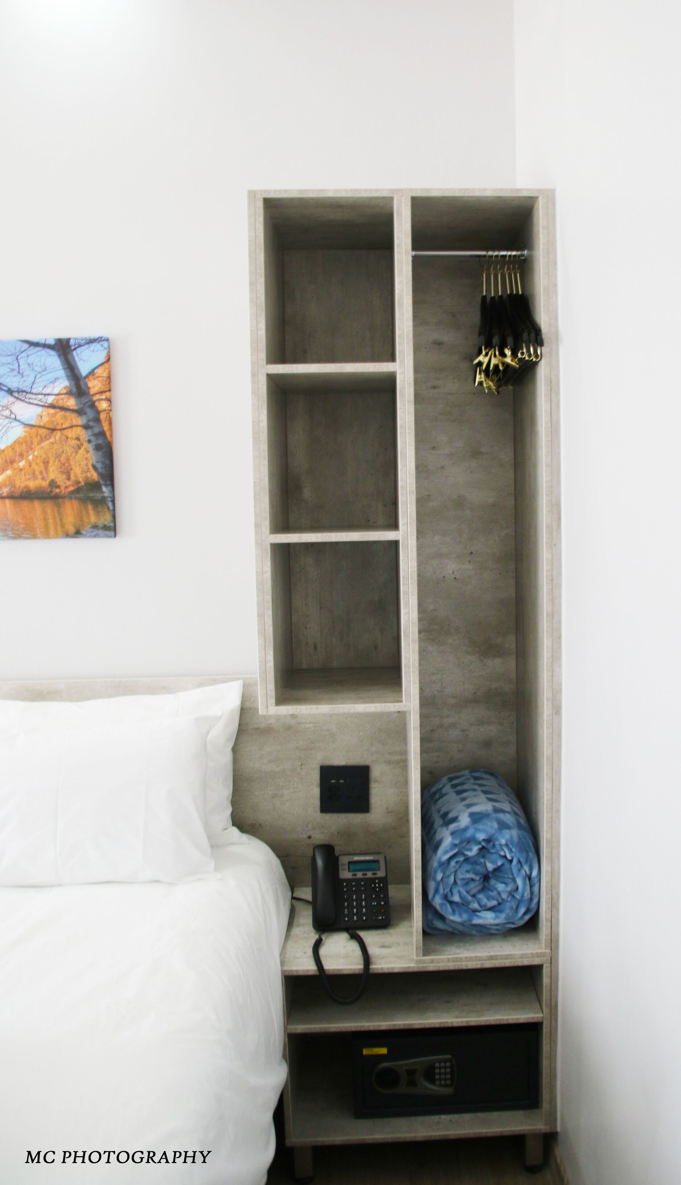 A hotel room with a bed, a wardrobe with hanging clothes, a phone, and a blanket.