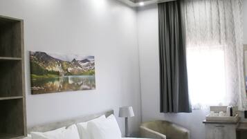 A hotel room with a bed, a desk, a chair, a painting, and a window with curtains.
