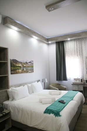 A hotel room with a bed, a desk, a chair, a painting, and a window with curtains.