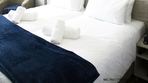 A hotel room with a neatly made bed, a painting of a waterfall, a chair, and a bedside table with a lamp.