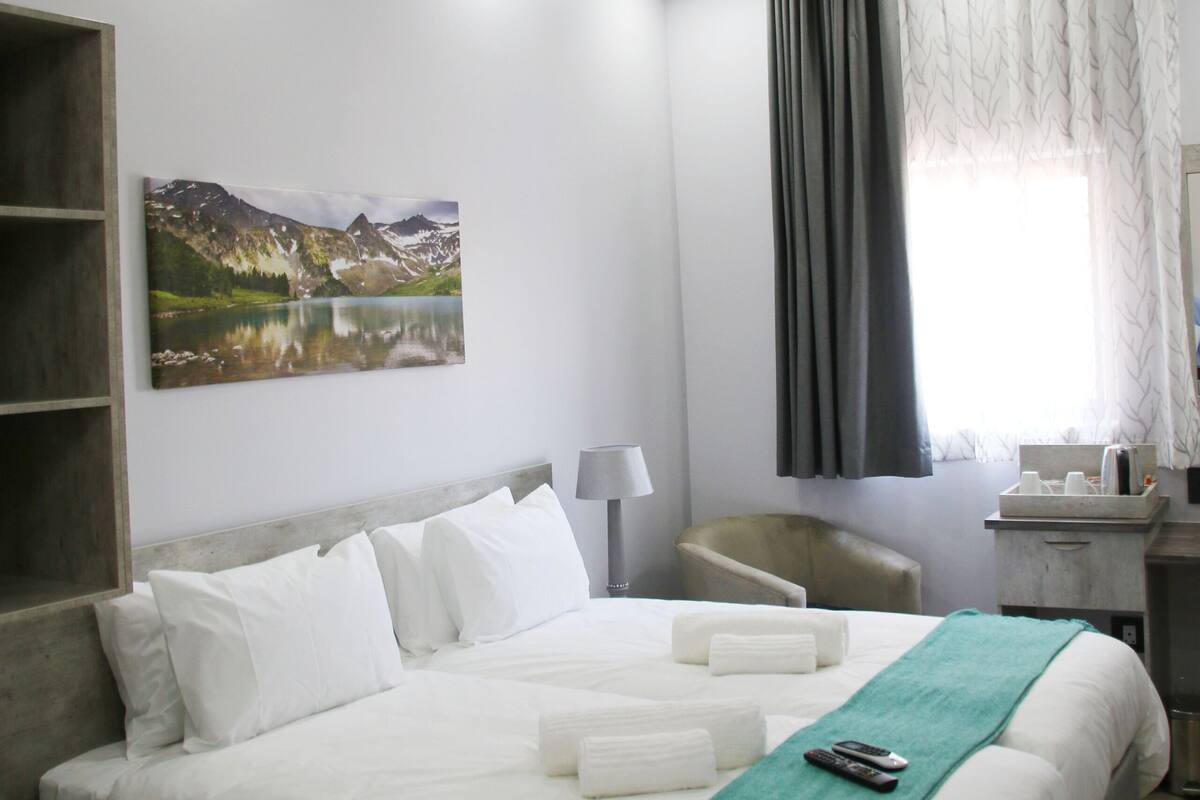 A hotel room with a bed, a desk, a chair, a painting, and a window with curtains.