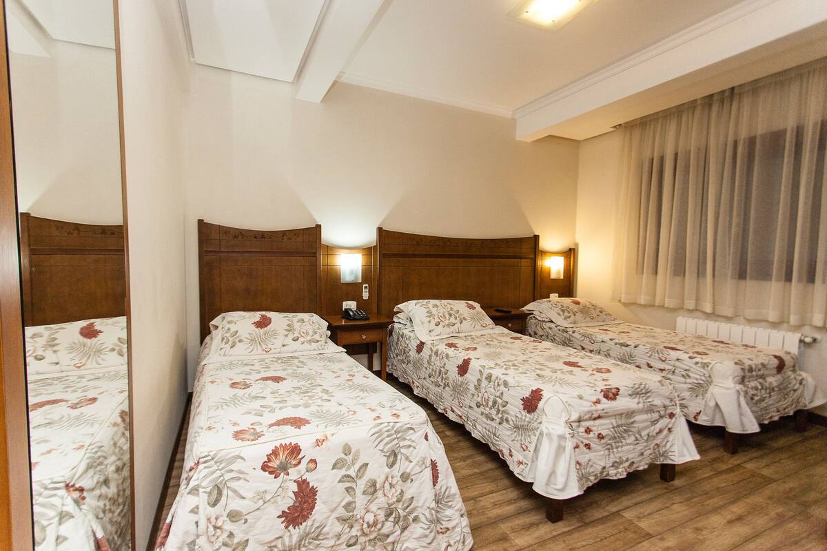 A hotel room with three single beds, wooden headboards, bedside tables, and a large mirror.