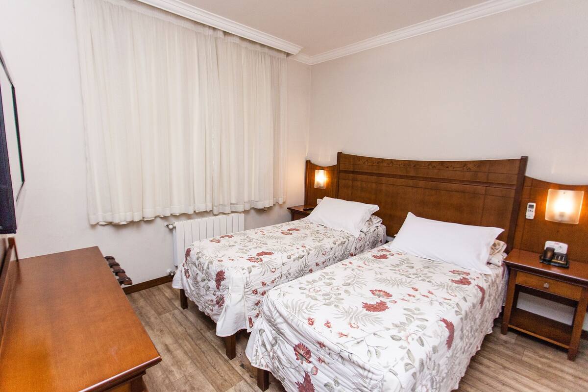 A hotel room with two single beds, a wooden nightstand with a lamp, a wooden desk, and a window with curtains.