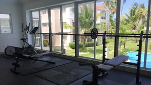 A gym with exercise equipment, large windows, and a view of a swimming pool and greenery. - Punta Cana Corte Sea (Punta Cana)