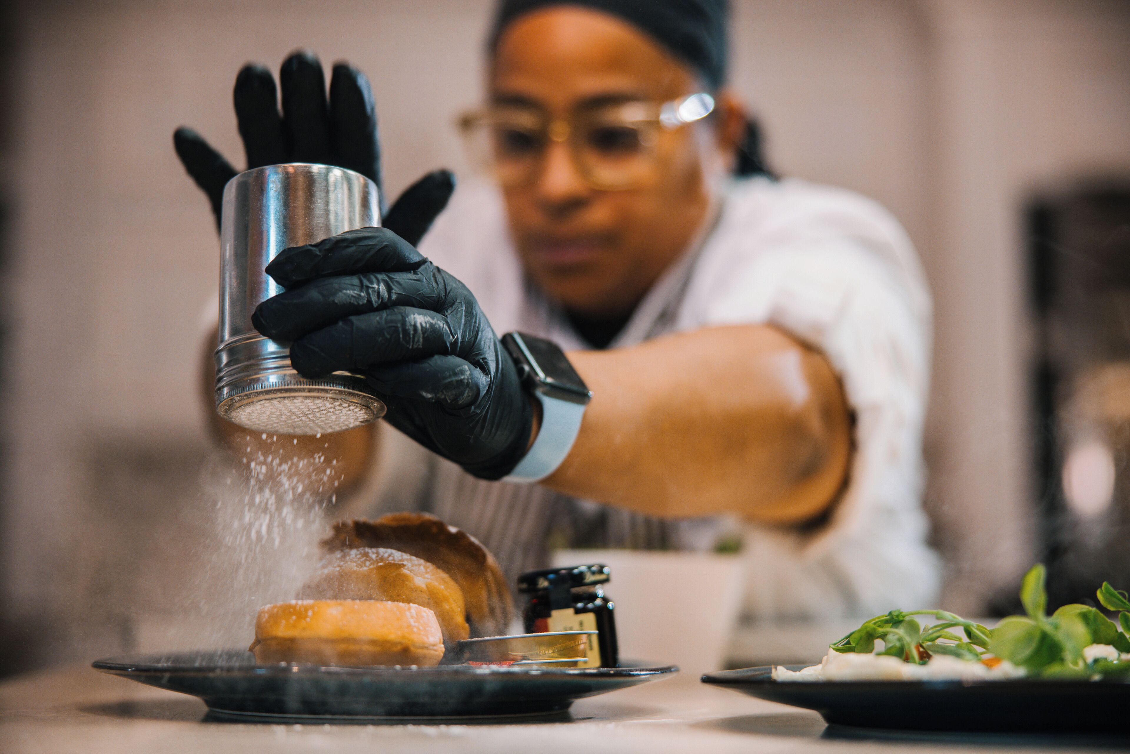 A person wearing black gloves handling a metal container over a plate with food, possibly seasoning it.