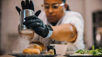 A person wearing black gloves handling a metal container over a plate with food, possibly seasoning it.