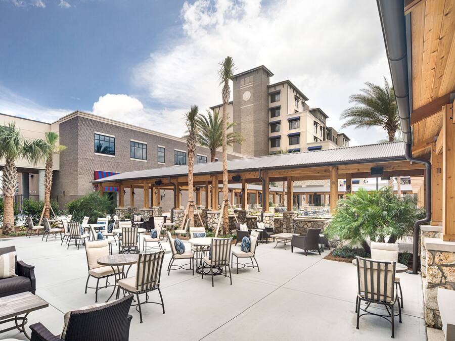 Un espace extérieur aménagé en terrasse, doté de plusieurs coins salon, comprenant des chaises et des tables, entouré de palmiers et bordé d’un bâtiment à la façade en briques.
