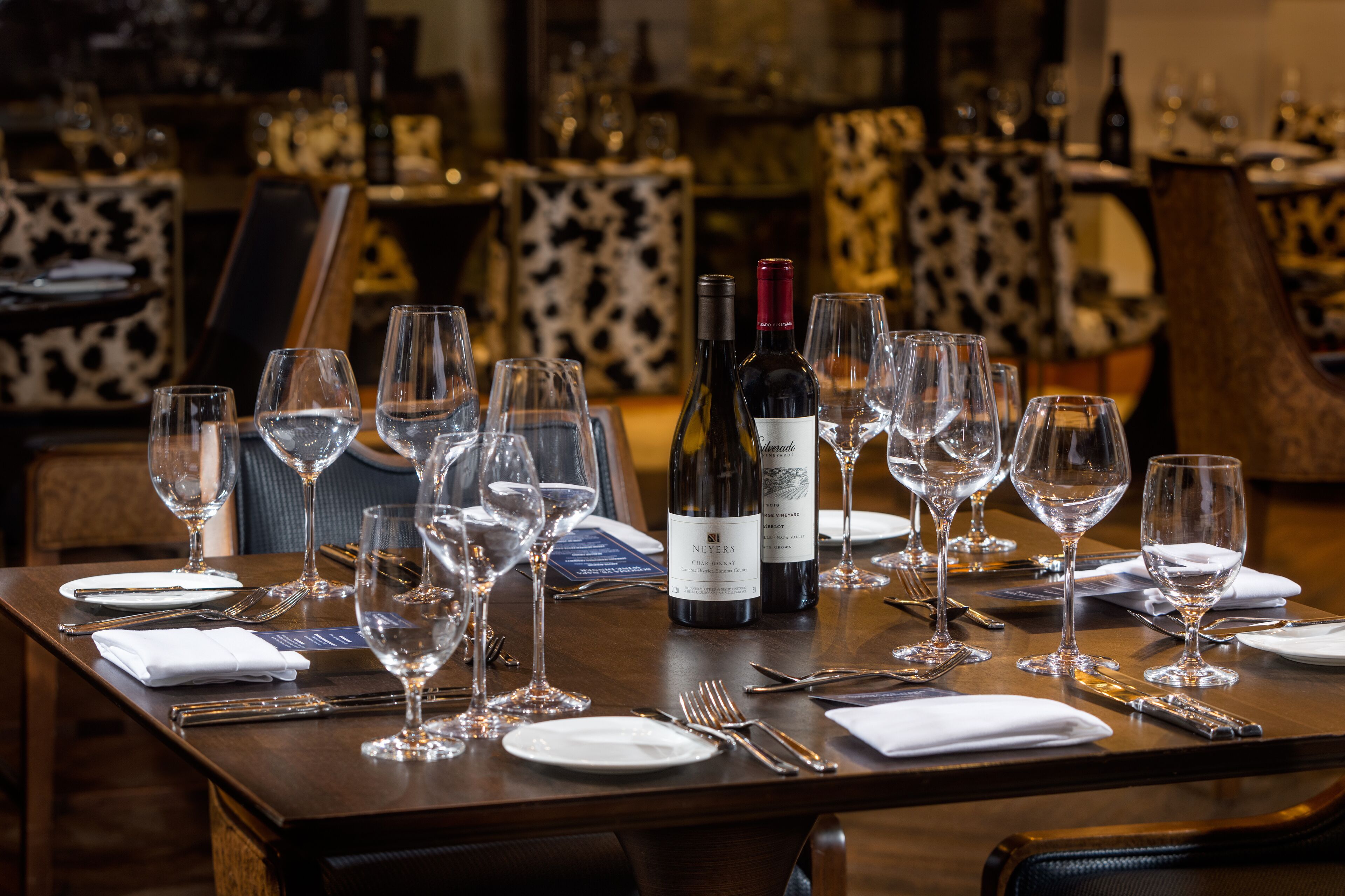 A dining table set with wine glasses, plates, and cutlery, with a bottle of wine and a menu.
