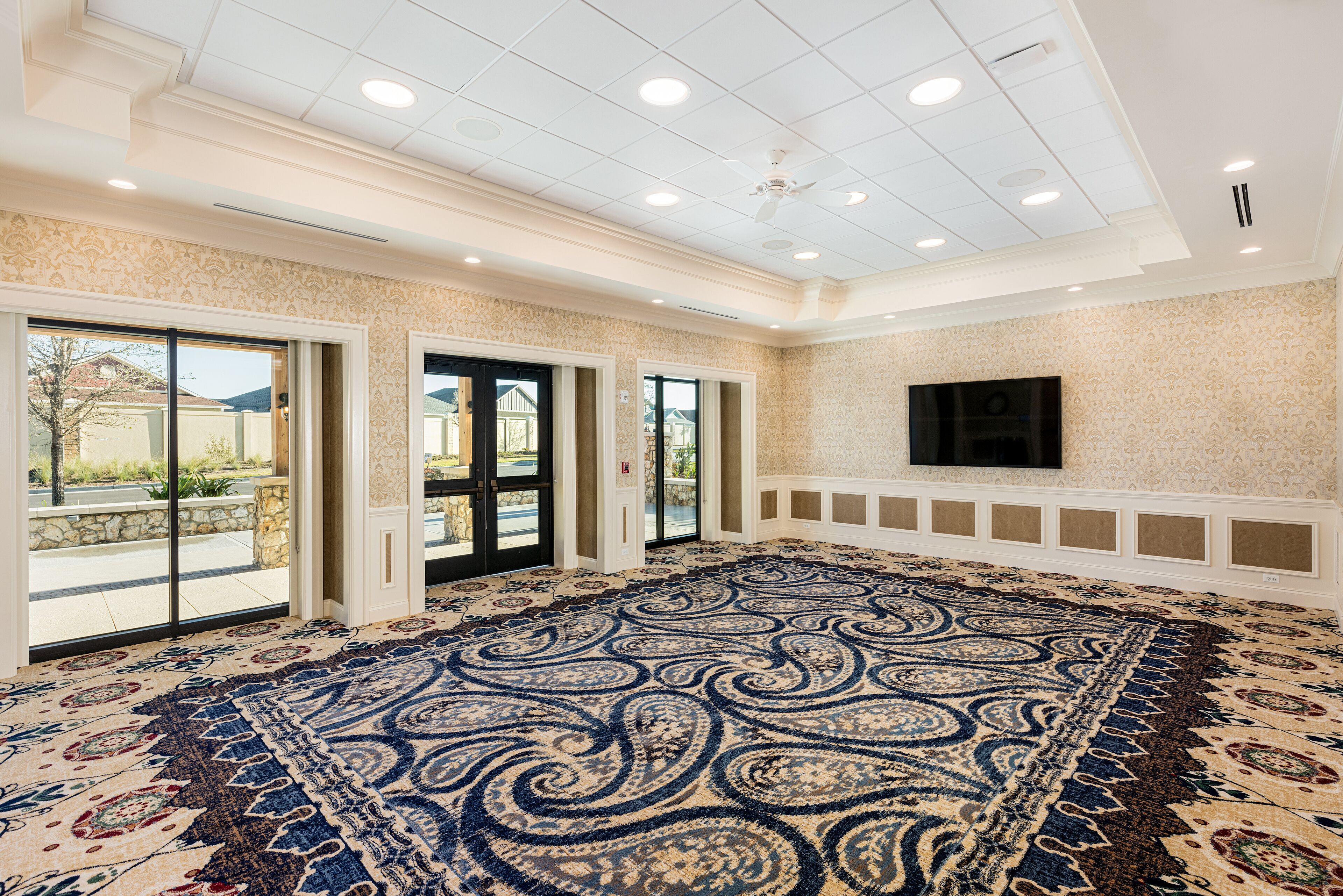 A spacious room with a large patterned carpet, a flat-screen TV mounted on the wall, and a ceiling fan.