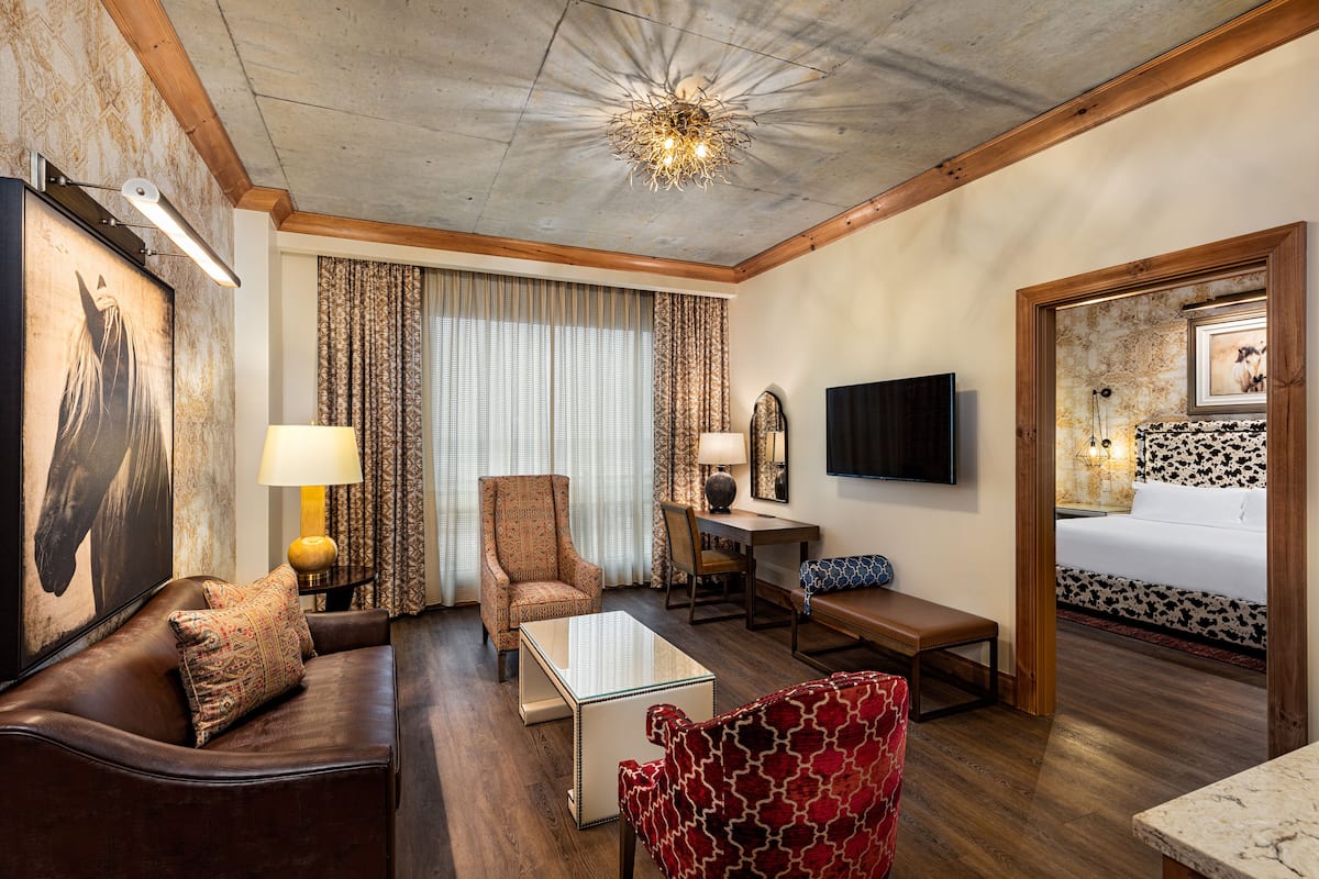A hotel room with a wooden ceiling, a large bed with a floral headboard, a brown leather sofa, a red patterned armchair, a glass coffee table, a wooden desk with a lamp, and a television on the wall.