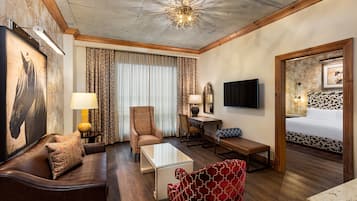Une chambre d’hôtel avec un plafond en bois, un grand lit doté d’une tête de lit à motifs floraux, un canapé en cuir marron, un fauteuil à motifs rouges, une table basse en verre, un bureau en bois avec une lampe et un téléviseur fixé au mur.
