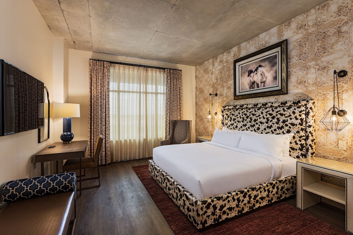 A hotel room with a large bed, a desk with a lamp, a chair, and a window with curtains.