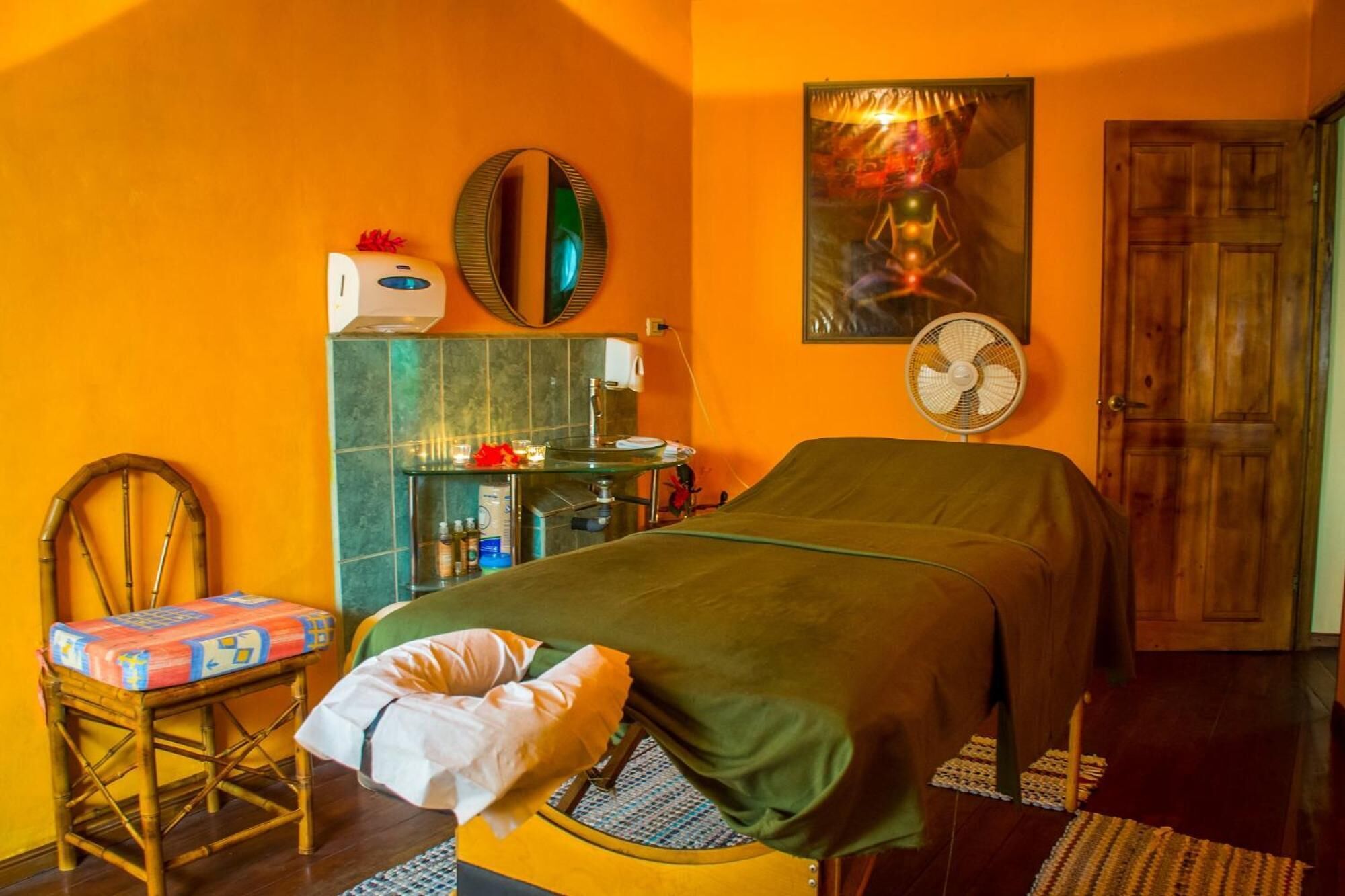 Quarto com cama, cadeira, espelho, ventilador e porta.