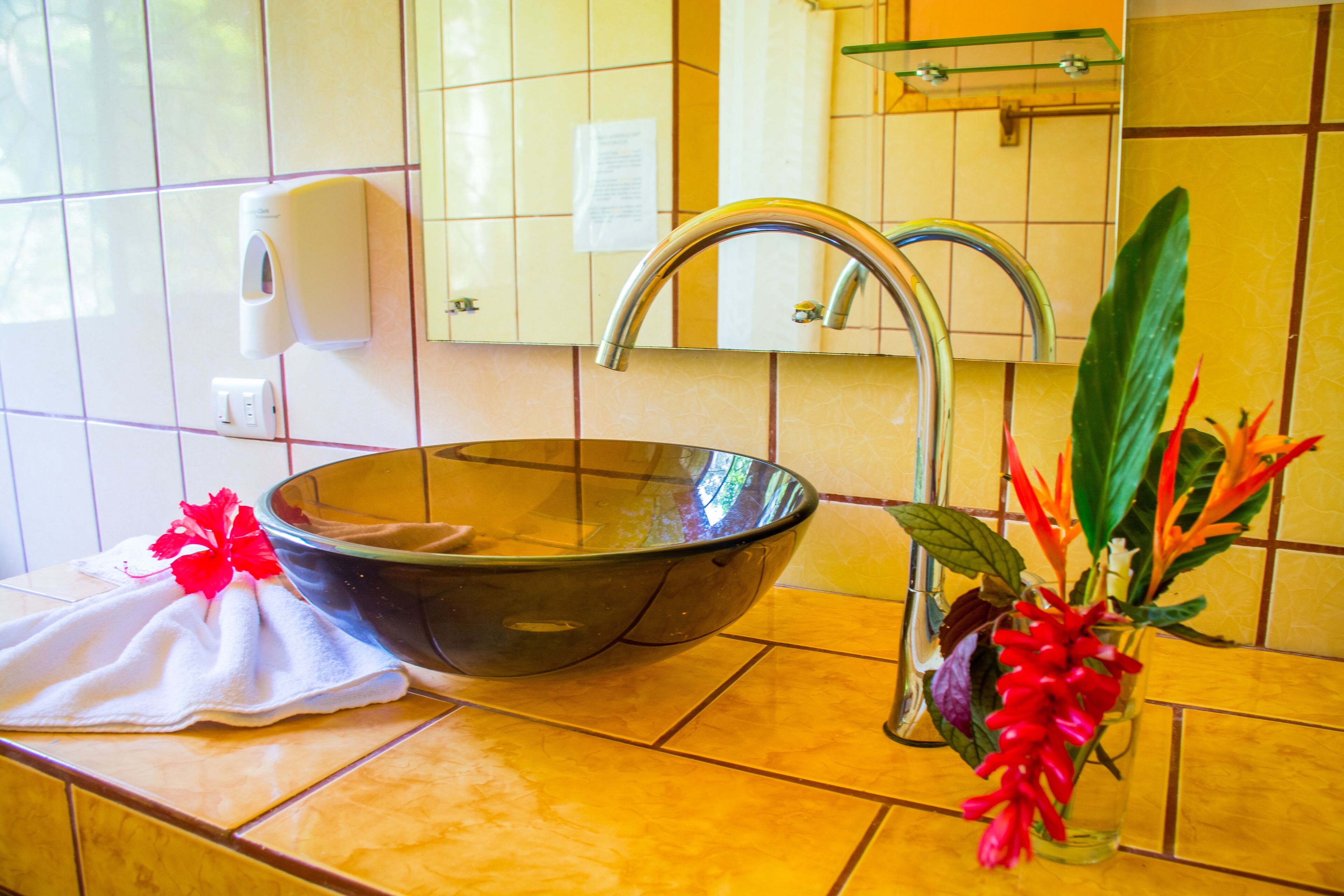 A bathroom with a large glass bowl sink, a towel, a soap dispenser, and a vase with flowers.