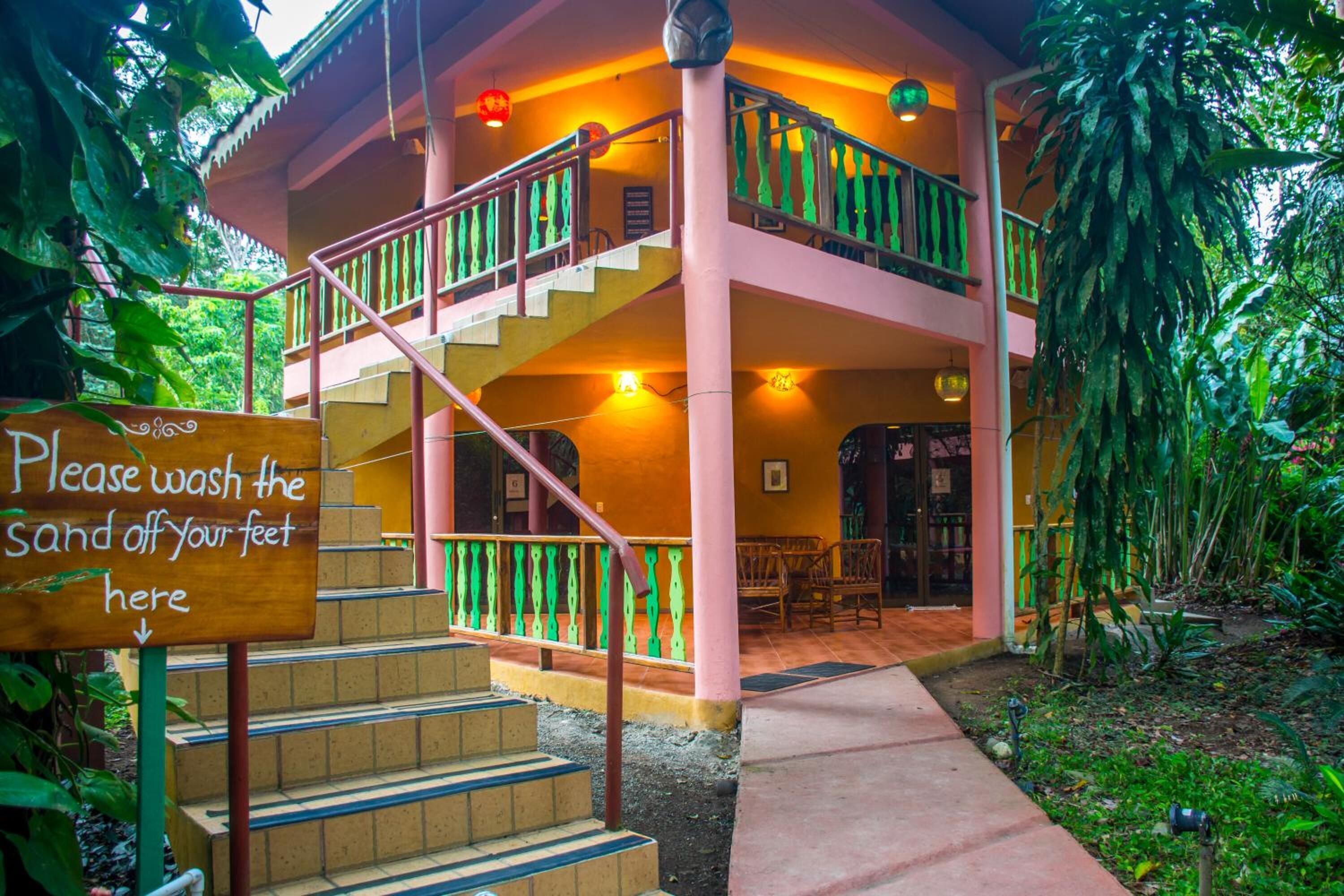 A two-story building with a staircase leading to a balcony, surrounded by lush greenery.