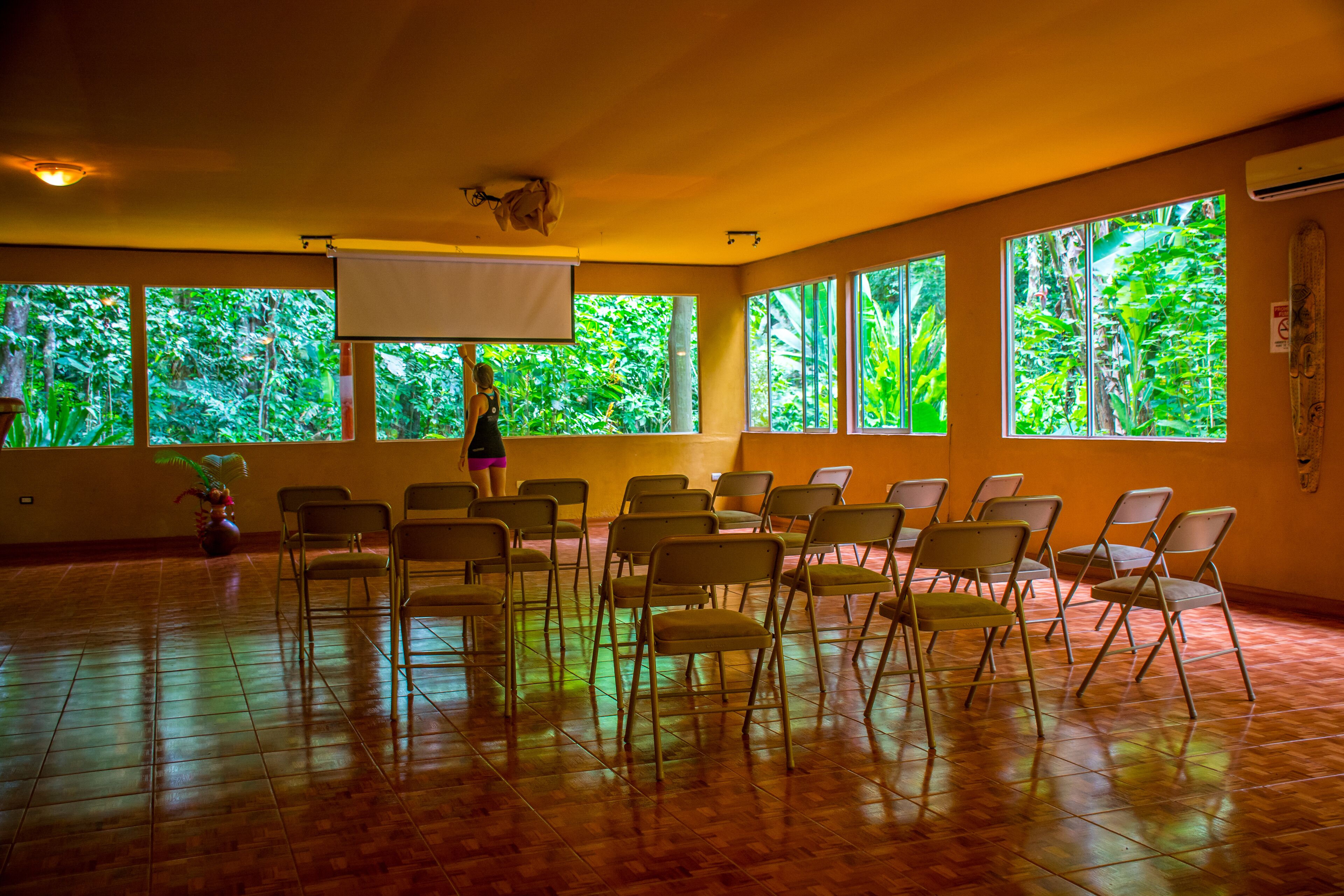 A room with large windows, chairs arranged in rows, and a projector screen.