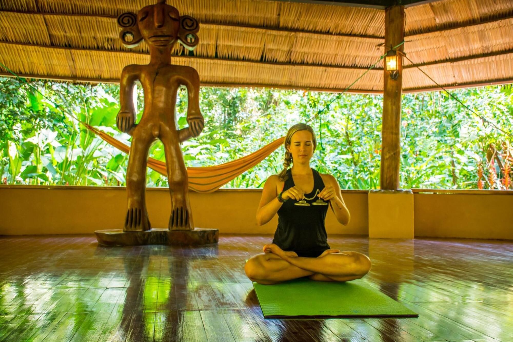 A person meditating on a yoga mat in a room with a thatched roof and a wooden sculpture.