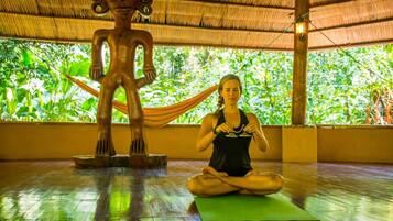 A person meditating on a yoga mat in a room with a thatched roof and a wooden sculpture.