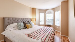 Un dormitorio con cabecera capitonada, una cama grande con ropa de cama blanca, almohadas estampadas y una manta colorida. Hay dos ventanas con persianas, una de ellas con un adorno de abanico.