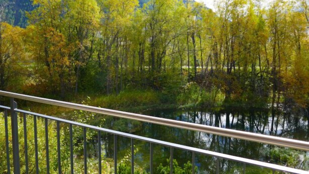 A view of a river with trees reflecting on the water, a mountain in the background, and a metal railing in the foreground.