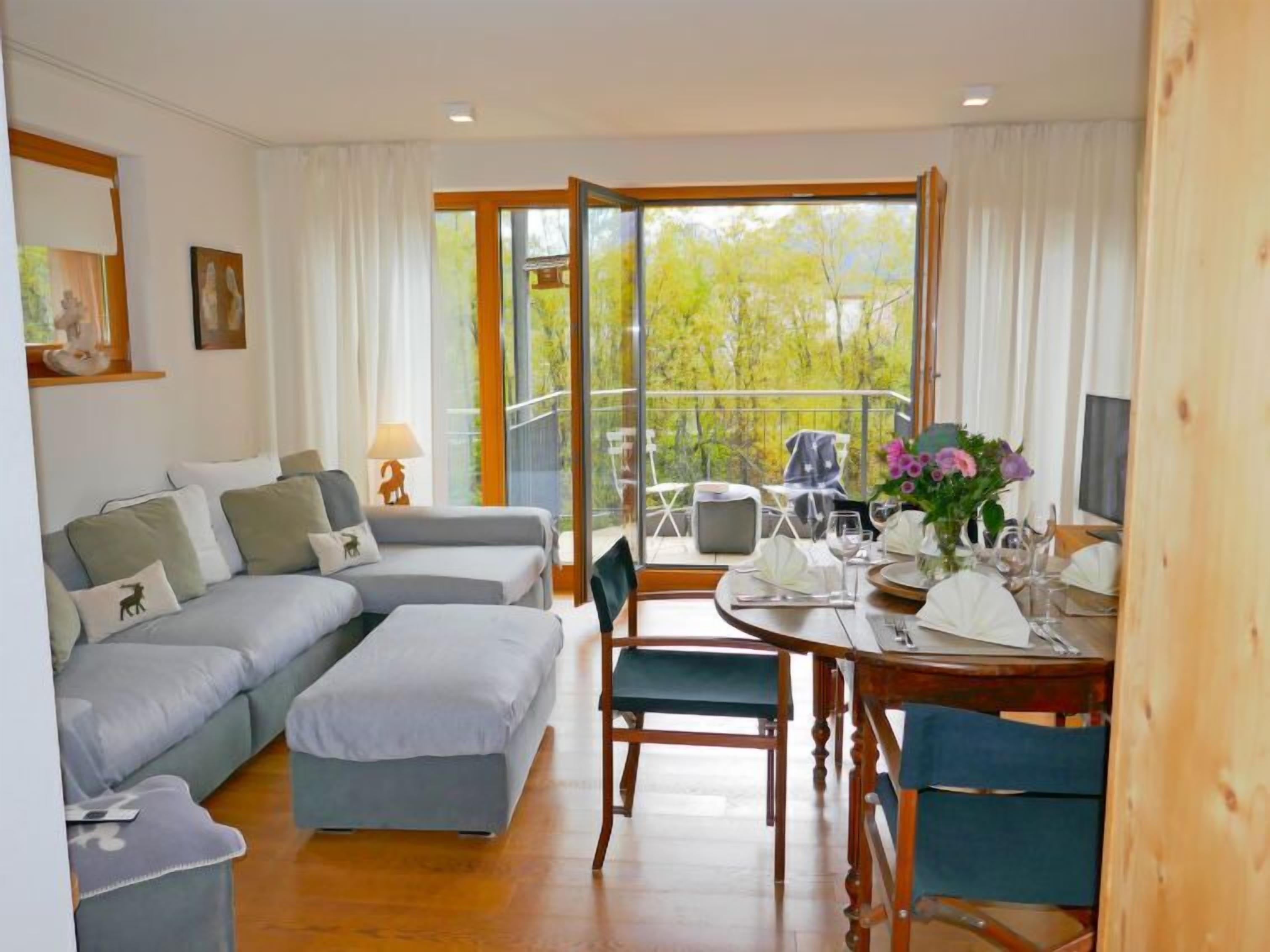 A modern living room with a sofa, a dining table set for two, and a balcony with a view of trees.