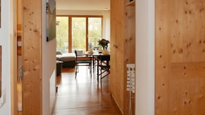 A wooden interior hallway leading to a dining area with a table and chairs, large windows, and a sofa.