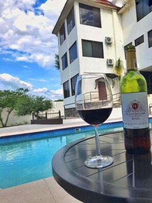 A glass of red wine next to a bottle labeled "El Candelero Hotel" by a pool with a modern building in the background. - Hotel Candelero (Yuriria)