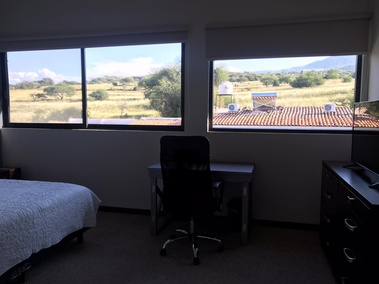 Un dormitorio con cama, escritorio y silla, con ventanas amplias que ofrecen vistas a un paisaje rural.