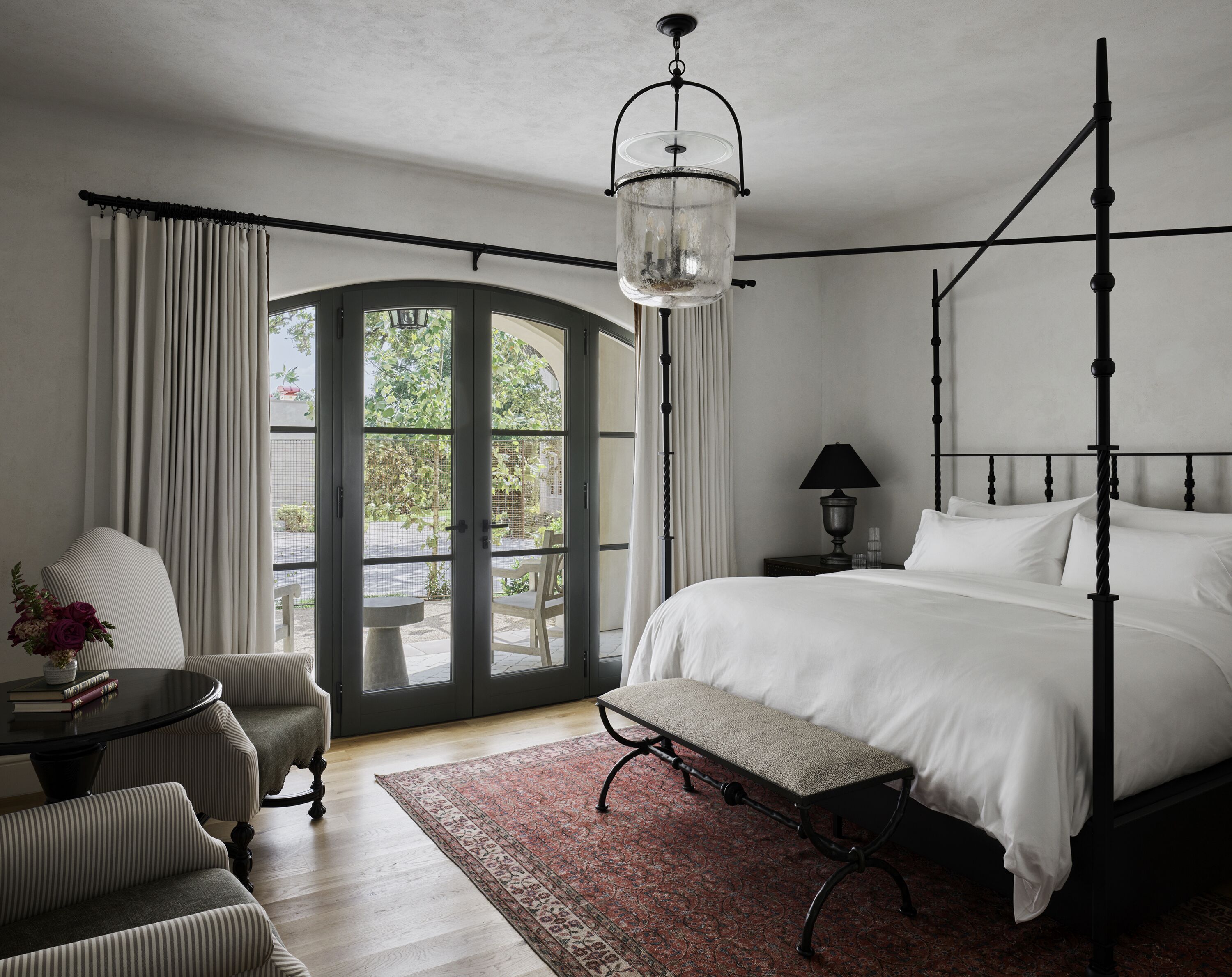 A bedroom with a four-poster bed, a bench, a table with chairs, and a view through a glass door.