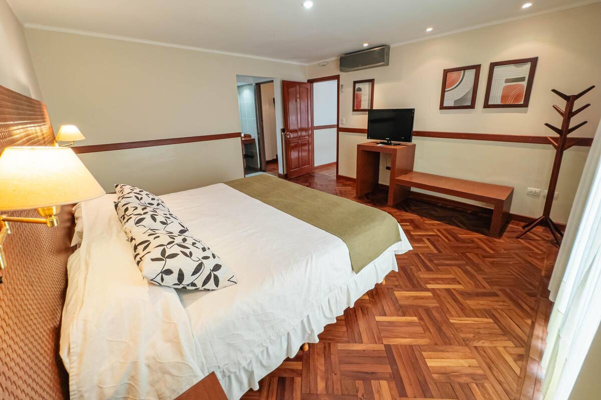 Quarto de hotel com cama, luminária de cabeceira, televisão, um banco e piso de madeira.