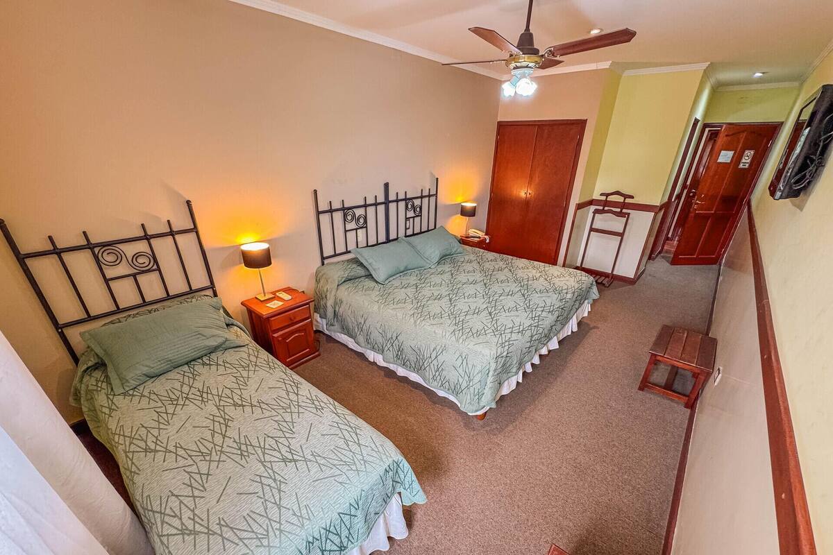 Quarto com duas camas, um ventilador de teto, mesa de cabeceira com abajur e um banquinho de madeira.
