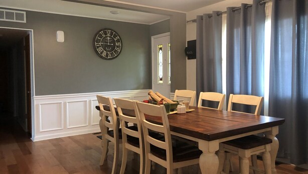Une salle à manger avec une table en bois et des chaises blanches, une horloge au mur et une fenêtre avec des rideaux.